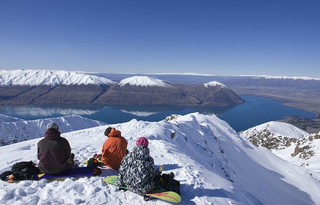 Three people sitting on their snowboards on top of Mount Ohau gaze out at the view of the lake below and the Southern Alps during a break on their NZ snow tour