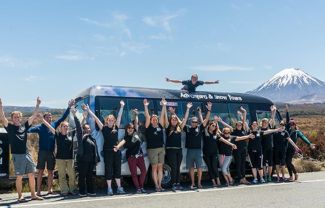 A group of people with their arms raised in joy stand in front of a New Zealand tour bus with a Mount Ngauruhoe in the distance